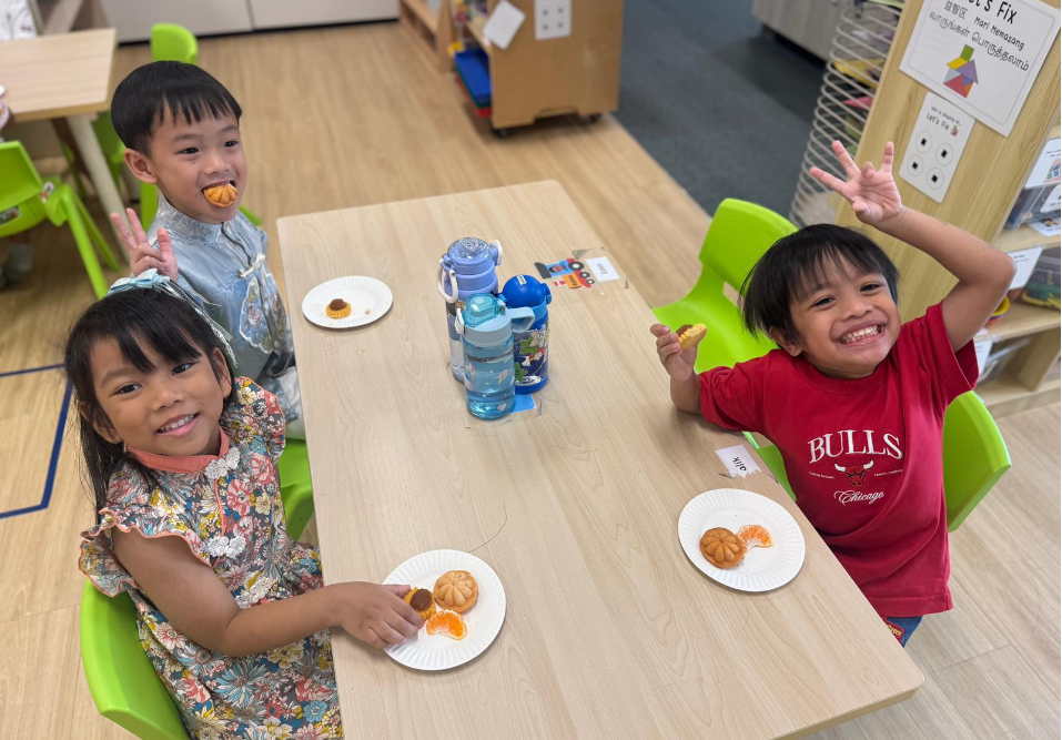 Three children smile at a classroom table with small snacks on paper plates and water bottles in the center.
